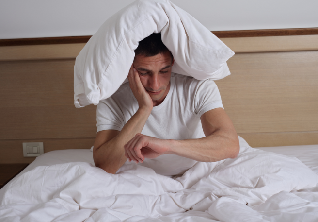 Mann mit weißem Kissen auf dem Kopf sitzt im Bett und schaut müde auf sein linkes Handgelenk.